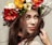 A woman is adorned with a vibrant floral crown featuring red, white, yellow, and orange flowers. Her long brown hair flows naturally around her face as she gazes softly at the viewer with a subtle smile. The background is a neutral color, highlighting the colorful flowers and her expressive features.
