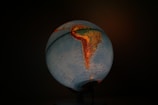 A globe illuminated from within, highlighting the continents of South America and parts of North America against a dark background. The map features geographic details with countries and major cities marked. Earth tones, such as blues and greens for oceans and landmasses, dominate the image.
