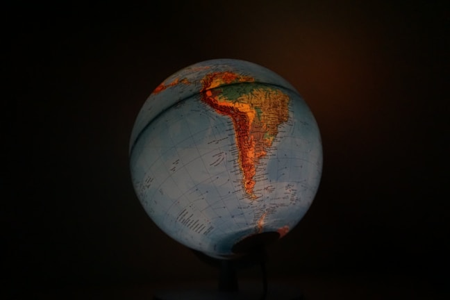 A globe illuminated from within, highlighting the continents of South America and parts of North America against a dark background. The map features geographic details with countries and major cities marked. Earth tones, such as blues and greens for oceans and landmasses, dominate the image.