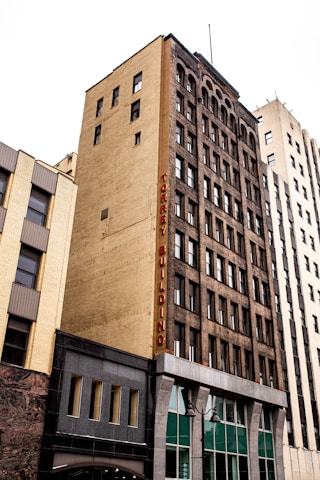 A tall, multi-story urban building with a yellow brick facade and dark window frames, featuring a vertical sign that reads 'TORREY BUILDING' in red letters. The building stands alongside other high-rise structures in a city setting.