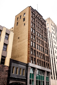 A tall, multi-story urban building with a yellow brick facade and dark window frames, featuring a vertical sign that reads 'TORREY BUILDING' in red letters. The building stands alongside other high-rise structures in a city setting.
