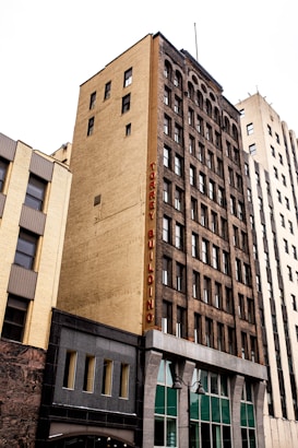 A tall, multi-story urban building with a yellow brick facade and dark window frames, featuring a vertical sign that reads 'TORREY BUILDING' in red letters. The building stands alongside other high-rise structures in a city setting.
