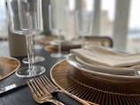 A close-up of a dining table setting features elegant gold-rimmed plates stacked on top of each other, topped with a neatly folded napkin. Two gold forks are placed beside the plates. Crystal wine glasses stand tall in the background, and golden placemats enhance the luxurious feel of the setting.