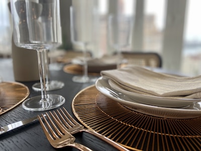 A close-up of a dining table setting features elegant gold-rimmed plates stacked on top of each other, topped with a neatly folded napkin. Two gold forks are placed beside the plates. Crystal wine glasses stand tall in the background, and golden placemats enhance the luxurious feel of the setting.