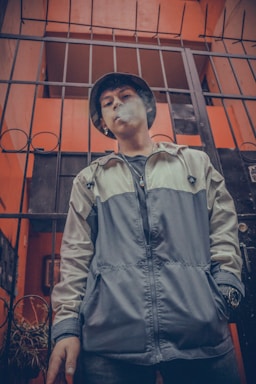 A person stands in front of a metal gate, blowing smoke. The individual is wearing a bucket hat and a two-tone jacket. The background includes an orange wall and part of a building interior.