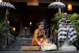 A woman dressed in traditional Balinese attire arranges a ceremonial offering on a stone step in front of a restaurant entrance. She is surrounded by plants and an intricately dressed stone statue. Two umbrellas with checkered patterns flank the pathway, and a wooden sign with the text 'Uma Resto' is seen above.