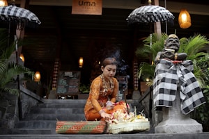 A woman dressed in traditional Balinese attire arranges a ceremonial offering on a stone step in front of a restaurant entrance. She is surrounded by plants and an intricately dressed stone statue. Two umbrellas with checkered patterns flank the pathway, and a wooden sign with the text 'Uma Resto' is seen above.