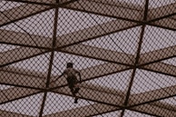 A person climbs a large geometric metal structure made of intersecting beams, creating a diamond pattern. The scene is silhouetted against a muted sky, giving an impression of adventure and exploration.