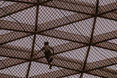 A person climbs a large geometric metal structure made of intersecting beams, creating a diamond pattern. The scene is silhouetted against a muted sky, giving an impression of adventure and exploration.