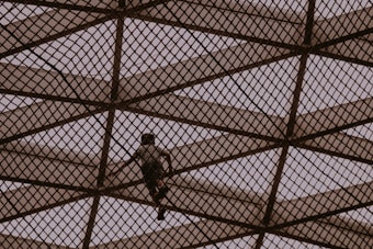 A person climbs a large geometric metal structure made of intersecting beams, creating a diamond pattern. The scene is silhouetted against a muted sky, giving an impression of adventure and exploration.