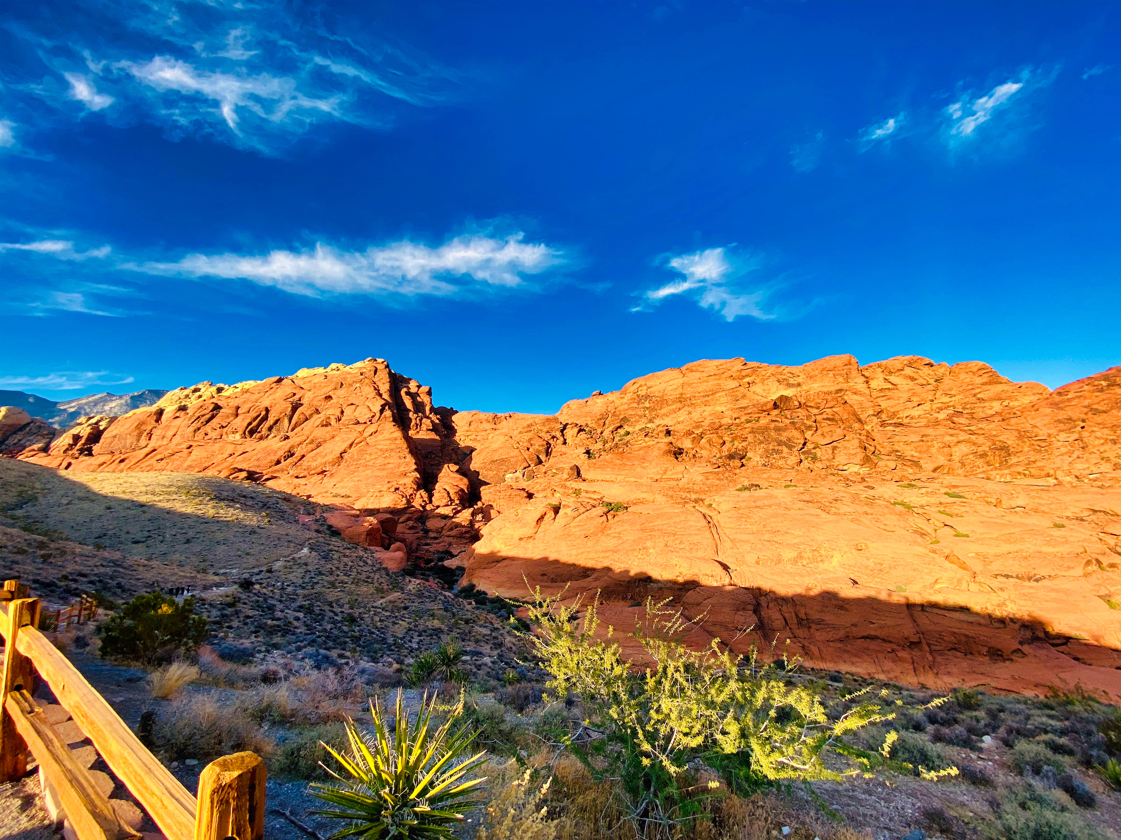 Glowing red sandstone cliffs at Red Rock Canyon lit by golden-hour sunlight, with a wooden trail fence in the foreground.