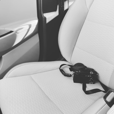 A black and white camera rests on a light-colored car seat with a seatbelt. The car door and part of the window frame are visible, casting shadows inside the vehicle.
