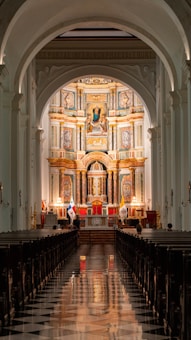A grand and intricately designed church interior with a long, reflective marble aisle leading towards an ornate altar. The altar is adorned with religious iconography, gold accents, and flags. Tall, white arches frame the space, enhancing the vertical splendor of the architecture.