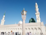 A mosque with a large green dome and several minarets against a clear blue sky. A variety of people in diverse attire are walking and standing in the open courtyard.