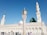 A mosque with a large green dome and several minarets against a clear blue sky. A variety of people in diverse attire are walking and standing in the open courtyard.