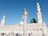 A mosque with a large green dome and several minarets against a clear blue sky. A variety of people in diverse attire are walking and standing in the open courtyard.