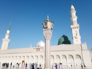 A mosque with a large green dome and several minarets against a clear blue sky. A variety of people in diverse attire are walking and standing in the open courtyard.