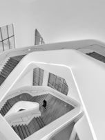 A modern architectural interior featuring multiple levels and sleek, curved staircases. The clean lines and open spaces create a minimalist and futuristic ambiance. One person is visible walking through the structure, adding a sense of scale to the design.