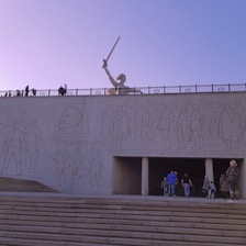 A monumental structure features a large sculpture of a figure holding a sword above a wall with engraved illustrations of people. The scene depicts historical or commemorative imagery with a strong cultural or nationalistic tone. People are seen walking near the steps that lead to the wall.
