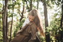 A person with long brown hair leans forward slightly in a peaceful, sun-dappled forest. The trees are lush with green foliage and sunlight filters through the branches, creating a serene atmosphere. The person is wearing an earth-toned coat, enhancing the natural setting.