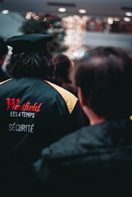 People are standing in what appears to be a shopping center or mall. In the foreground, there is a person wearing a security uniform with 'Westfield Les 4 Temps' written on the back. The background is softly blurred with bokeh lights, suggesting a festive or decorated environment.