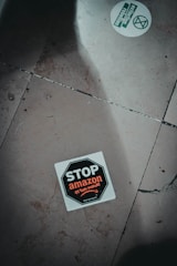 A sticker on the ground with the text 'STOP amazon et son monde' featuring a stop sign design. The sticker is positioned slightly off-center on a tiled floor which has visible cracks and dirt. Another round sticker with green text and graphics, related to activism, is partially visible.