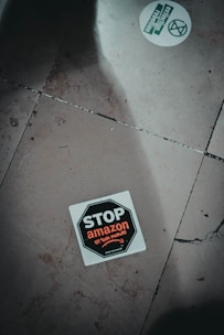 A sticker on the ground with the text 'STOP amazon et son monde' featuring a stop sign design. The sticker is positioned slightly off-center on a tiled floor which has visible cracks and dirt. Another round sticker with green text and graphics, related to activism, is partially visible.