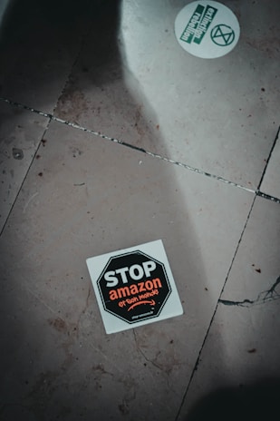 A sticker on the ground with the text 'STOP amazon et son monde' featuring a stop sign design. The sticker is positioned slightly off-center on a tiled floor which has visible cracks and dirt. Another round sticker with green text and graphics, related to activism, is partially visible.