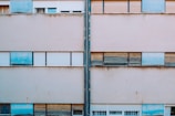 A concrete building wall with uniform rectangular windows, some of which have closed shutters. The wall is aged and shows signs of wear with varying tones of beige. Blue panels are present above and below some windows, contrasting with the muted tones.