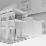 A detailed architectural model of a modern multi-story building, featuring clean lines and a minimalist design. The model showcases multiple floors with open balconies and large windows, emphasizing a contemporary architectural style displayed predominantly in white material.