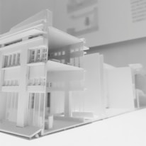 A detailed architectural model of a modern multi-story building, featuring clean lines and a minimalist design. The model showcases multiple floors with open balconies and large windows, emphasizing a contemporary architectural style displayed predominantly in white material.