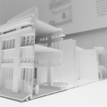 A detailed architectural model of a modern multi-story building, featuring clean lines and a minimalist design. The model showcases multiple floors with open balconies and large windows, emphasizing a contemporary architectural style displayed predominantly in white material.