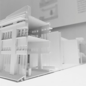 A detailed architectural model of a modern multi-story building, featuring clean lines and a minimalist design. The model showcases multiple floors with open balconies and large windows, emphasizing a contemporary architectural style displayed predominantly in white material.
