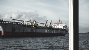 A large cargo ship with cranes on board, identified with the name 'HYUNDA' prominently displayed on its side, is navigating through a body of water. The sky is overcast, with clouds creating a subdued and slightly dramatic backdrop.