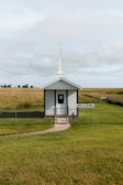 A small white chapel with a tall steeple sits in the middle of a grassy field, surrounded by a simple chain-link fence. A welcoming sign is displayed at the entrance. The sky above is overcast with soft clouds.