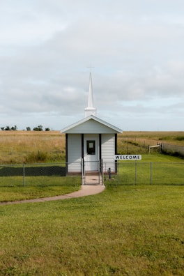 A small white chapel with a tall steeple sits in the middle of a grassy field, surrounded by a simple chain-link fence. A welcoming sign is displayed at the entrance. The sky above is overcast with soft clouds.