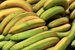 A large pile of ripe and unripe bananas, displaying various shades of yellow and green. The bananas are closely stacked, with some showing minor blemishes and spots.