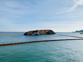 A partially submerged shipwreck is surrounded by calm blue waters, with containment booms floating around it. The sky is clear with few clouds, creating a serene backdrop. In the distance, a silhouette of land and a few structures can be seen.