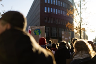 A crowd of people gathered outdoors, walking along a street. Some individuals are holding up protest signs, one of which prominently features an illustration of Earth in a trash can with the words 'Don't Trash It'. The scene is lit by a bright sun, casting long shadows. A tall building and trees with autumn leaves border the street.