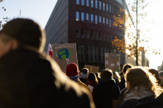 A crowd of people gathered outdoors, walking along a street. Some individuals are holding up protest signs, one of which prominently features an illustration of Earth in a trash can with the words 'Don't Trash It'. The scene is lit by a bright sun, casting long shadows. A tall building and trees with autumn leaves border the street.