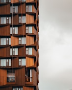 A modern building with a unique architectural design featuring a pattern of geometric brown panels and large windows. Several levels have small balconies. Two workers are suspended by ropes, performing maintenance or cleaning.