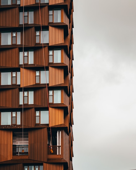 A modern building with a unique architectural design featuring a pattern of geometric brown panels and large windows. Several levels have small balconies. Two workers are suspended by ropes, performing maintenance or cleaning.
