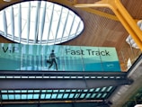 An interior view of a building featuring a sign for VIP Fast Track, with a graphic of a person carrying a suitcase. The ceiling has a modern design with wooden panels and a large circular skylight.