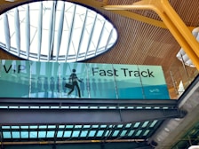 An interior view of a building featuring a sign for VIP Fast Track, with a graphic of a person carrying a suitcase. The ceiling has a modern design with wooden panels and a large circular skylight.