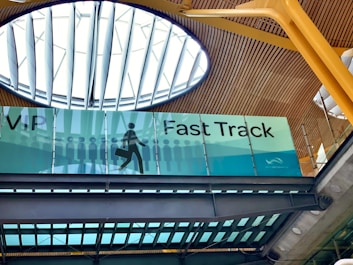 An interior view of a building featuring a sign for VIP Fast Track, with a graphic of a person carrying a suitcase. The ceiling has a modern design with wooden panels and a large circular skylight.