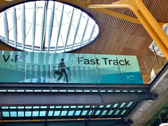 An interior view of a building featuring a sign for VIP Fast Track, with a graphic of a person carrying a suitcase. The ceiling has a modern design with wooden panels and a large circular skylight.