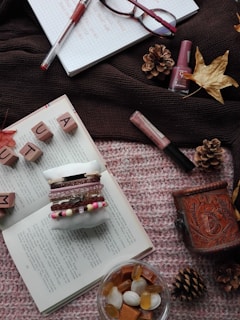 An autumnal-themed arrangement featuring a knitted brown scarf and an open book on a pink textured blanket. Wooden letter tiles spell out 'AUTUMN' while several pinecones and dried leaves enhance the seasonal vibe. A small glass dish contains a variety of candies, and a textured mug sits nearby. Several decorative bracelets are displayed on a fastened rolling pin, with makeup items such as nail polish and a lipstick placed around. A pair of glasses and a red pen rest on a checkered notebook.