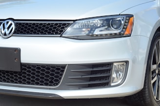 A close-up of the front portion of a white Volkswagen vehicle, showcasing the emblem, headlight, grille, and part of the bumper. The vehicle's design features a honeycomb grille and a sleek, modern headlight with LED elements.