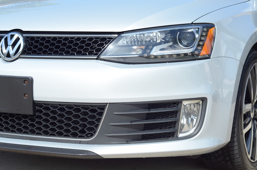 A close-up of the front portion of a white Volkswagen vehicle, showcasing the emblem, headlight, grille, and part of the bumper. The vehicle's design features a honeycomb grille and a sleek, modern headlight with LED elements.