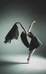 A dancer gracefully arches backward, balancing on one foot with long hair cascading down. She holds a flowing black fabric in one hand, creating a sense of motion. The background is softly lit, with subtle shadows accentuating her form.